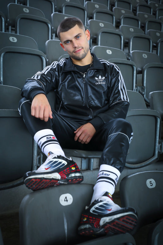 Model mit schwarzem glänzenden Adidas-Trainingsanzug, weißen Sk8erboy® Gay Sox Lightweight Crew Socks und Sneakern in Stadion-Sitzreihen – sportlicher Streetstyle-Look mit Fokus auf Socken-Statement.