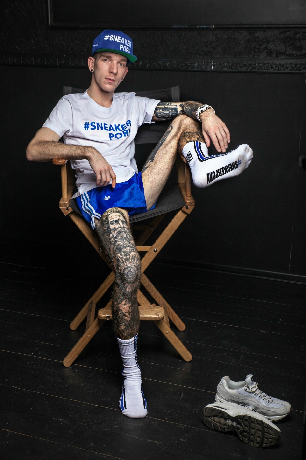 cooler junge mit adidas glanzshort und weissem sneakerporn t-shirt trägt sneakerporn socken in weiss und royalblau von sk8erboy sitzend im regiestuhl vor einer schwarzen wand