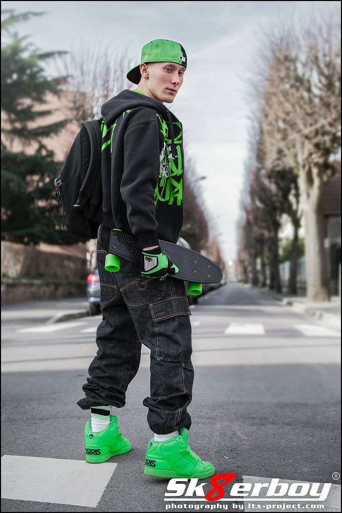 cooler skater boy mit kappe und rucksack trägt sein skateboard und baggy hose dazu grüne sneaker und passende sk8erboy deluxe socks in neon gruen