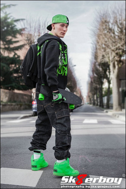 cooler skater boy mit kappe und rucksack trägt sein skateboard und baggy hose dazu grüne sneaker und passende sk8erboy deluxe socks in neon gruen