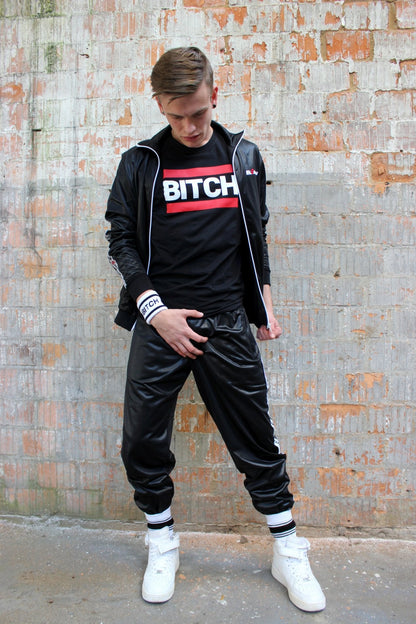 junge mit glanzanzug trainingsanzug von sk8erboy tragt ein schwarzes t-shirt mit bitch aufdruck und haelt sich den schritt mit blick auf seine weissen sneaker und socks von nike uns sk8erboy