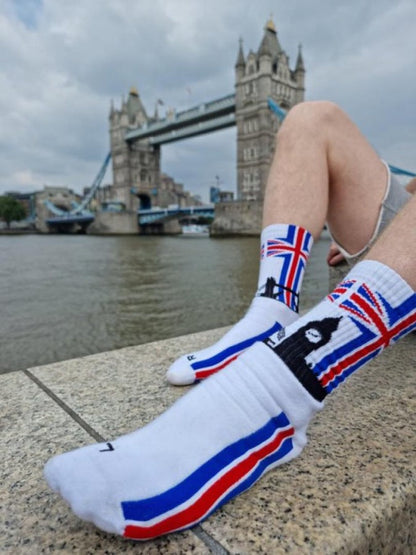 junger gay mit sk8erboy london socken mit union jack flagge am bund und big ben mit tower bridge mit extra grossem logo und schriftzug auf der unterseite der socken liegt vor der tower bridge