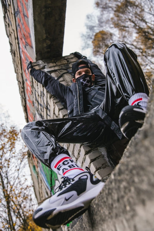 cooler gay mit pvc sporthose streckt sich an einer steinwand mit fck abl socks und nike air max sneaker im bild