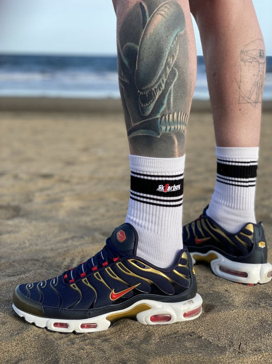 sk8erboy deluxe socken sox in weiss mit schwarzen ringen schwarzen mit schwarzem streifen und logo einstickung rote 8 acht er traegt nike tn in schwarz rot gold am strand mit meerblick und tattow am bein