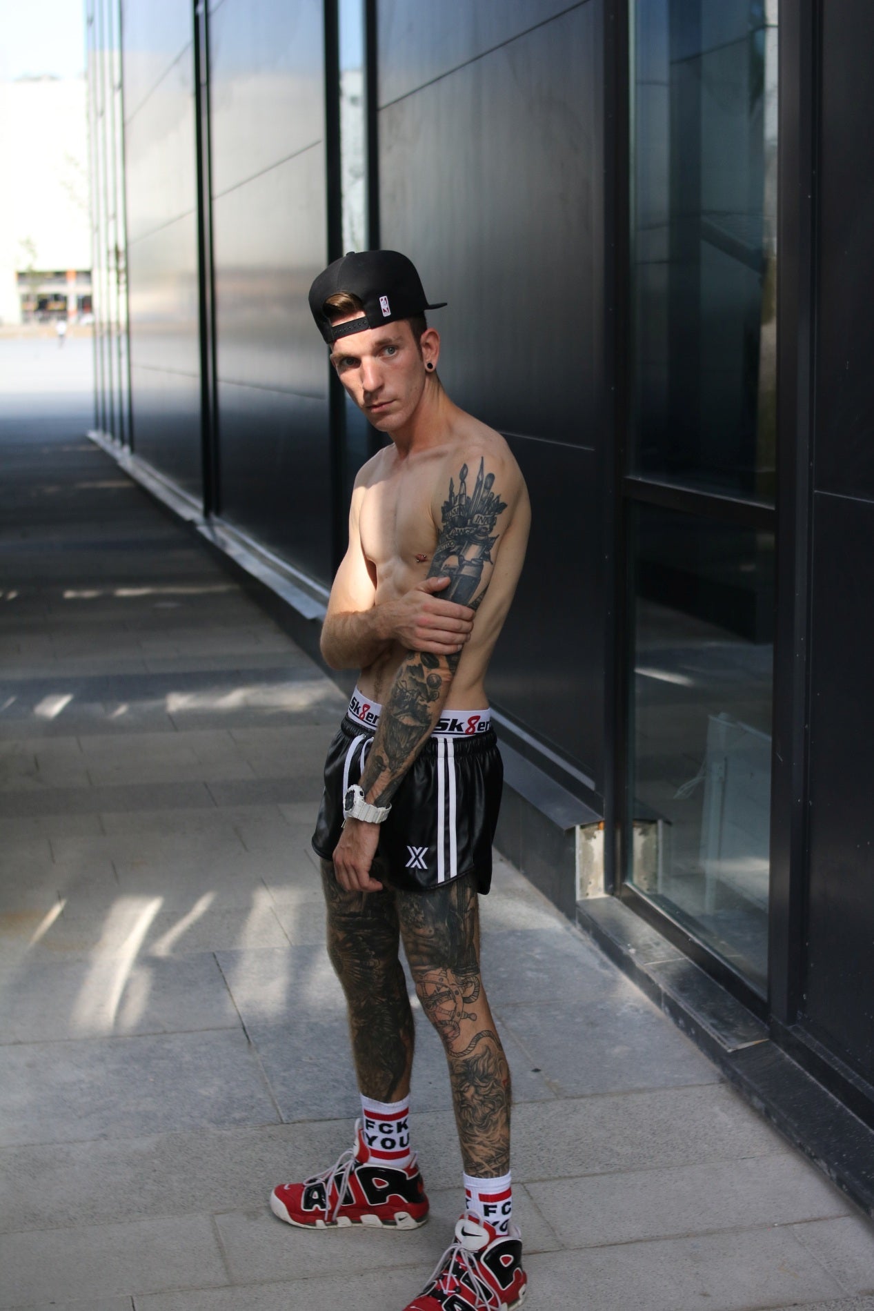 junger gay mit Tätowierungen am arm und beine von der seite mit fck you socks von sk8erboy und schwarzer boxer berlin short ueber dem jock mit roten nike air sneaker