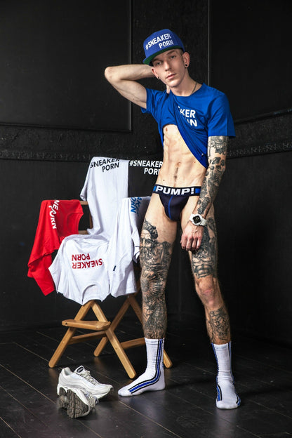 sportlicher junge zeigt seinen trainierten bauch unter einem blauen t-shirt mit jock von pump! und trägt dazu socken von sneakerporn in weiss und royalblau von sk8erboy in einem schwarzen zimmer neben einem regiestuhl der mit t-shirt in verschiedenen farben behängt ist