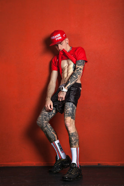 heißer junge mit tattoos am körper hat sein rotes shirt im mund und trägt einen jock von addicted mit sneakerporn socken von sk8erboy in weiss und feuerrot zu seinen schwarzen nike tn sneaker