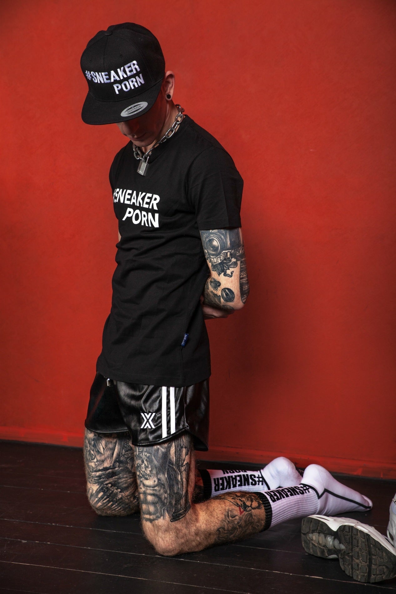 sportlicher typ kniet vor einer roten wand und trägt eine schwarze short von boxer berlin mit passenden socken von #sneakerporn by sk8erboy in weiss und schwarz mit schwarzem t-shirt und basecap