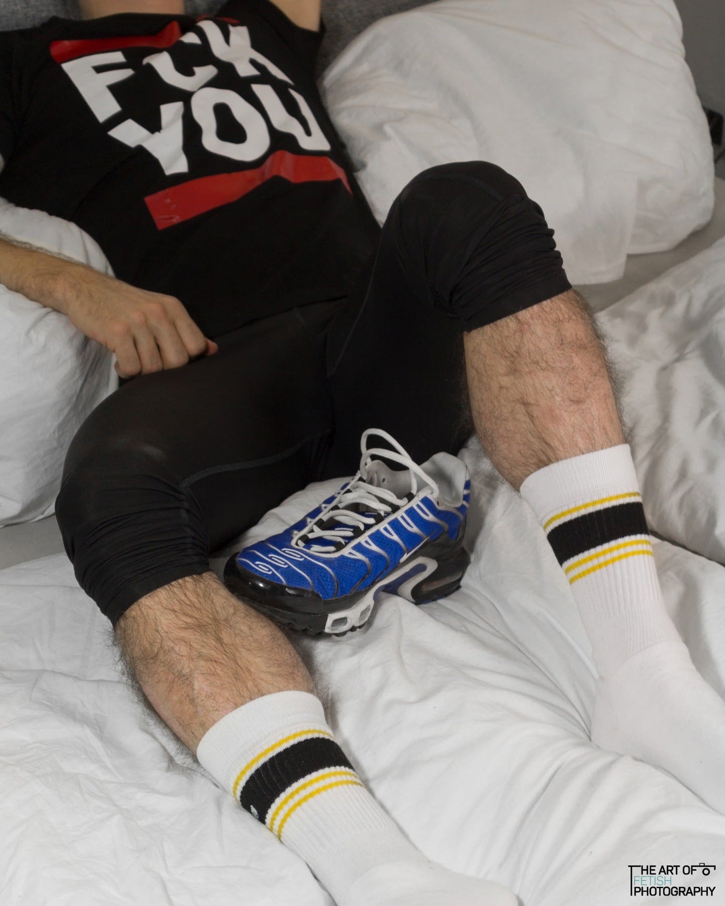 junge im bett liegend mit trainingshose und nike tn sneaker mit sk8erboy socken deluxe in gelb traegt ein schwarzes t-shirt mit dem aufdruck fck you in weiss mit roten balken