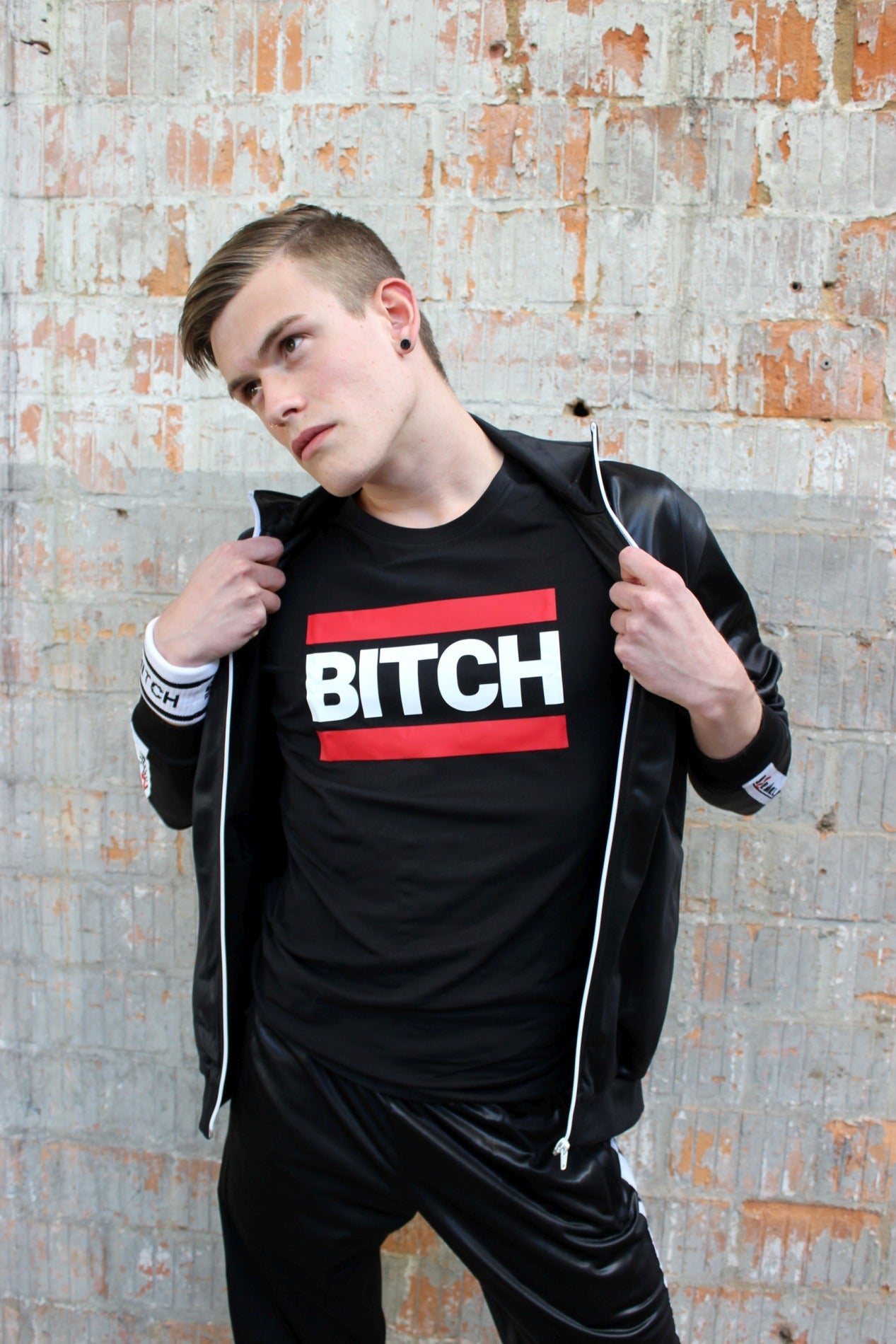 junge haelt seine glanzstoff jacke von sk8erboy geoeffnet und zeigt ein schwarzes t-shirt mit BITCH in großbuchstaben und rotem balken darueber und darunter