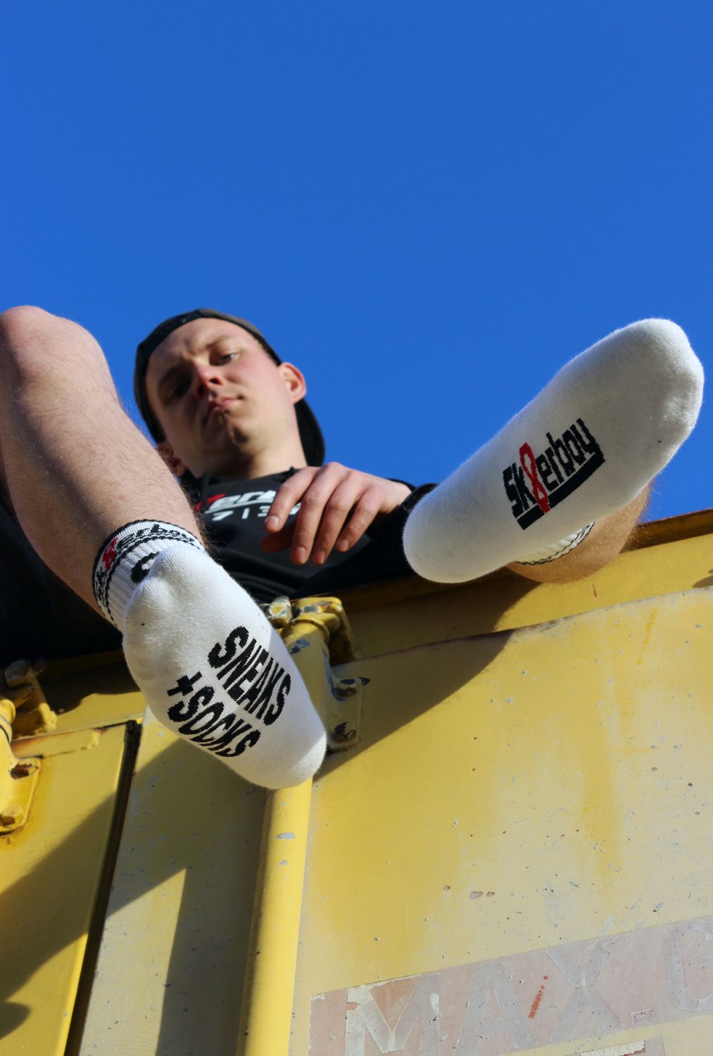 junger gay mit sneaks and socks socken von sk8erboy in weiß traegt einen schwarzen kaputzenpullover mi 365 7 24 aufdruck auf einem kontainer sitzend von unten 