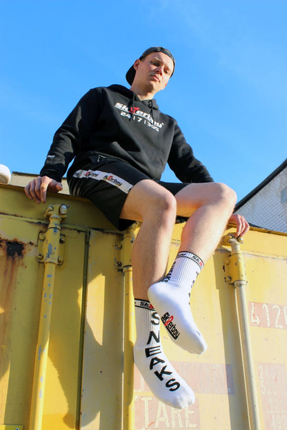 junger gay mit sneaks and socks socken von sk8erboy in weiß traegt einen schwarzen kaputzenpullover mi 365 7 24 aufdruck auf einem kontainer sitzend mit shiny short von sk8erboy