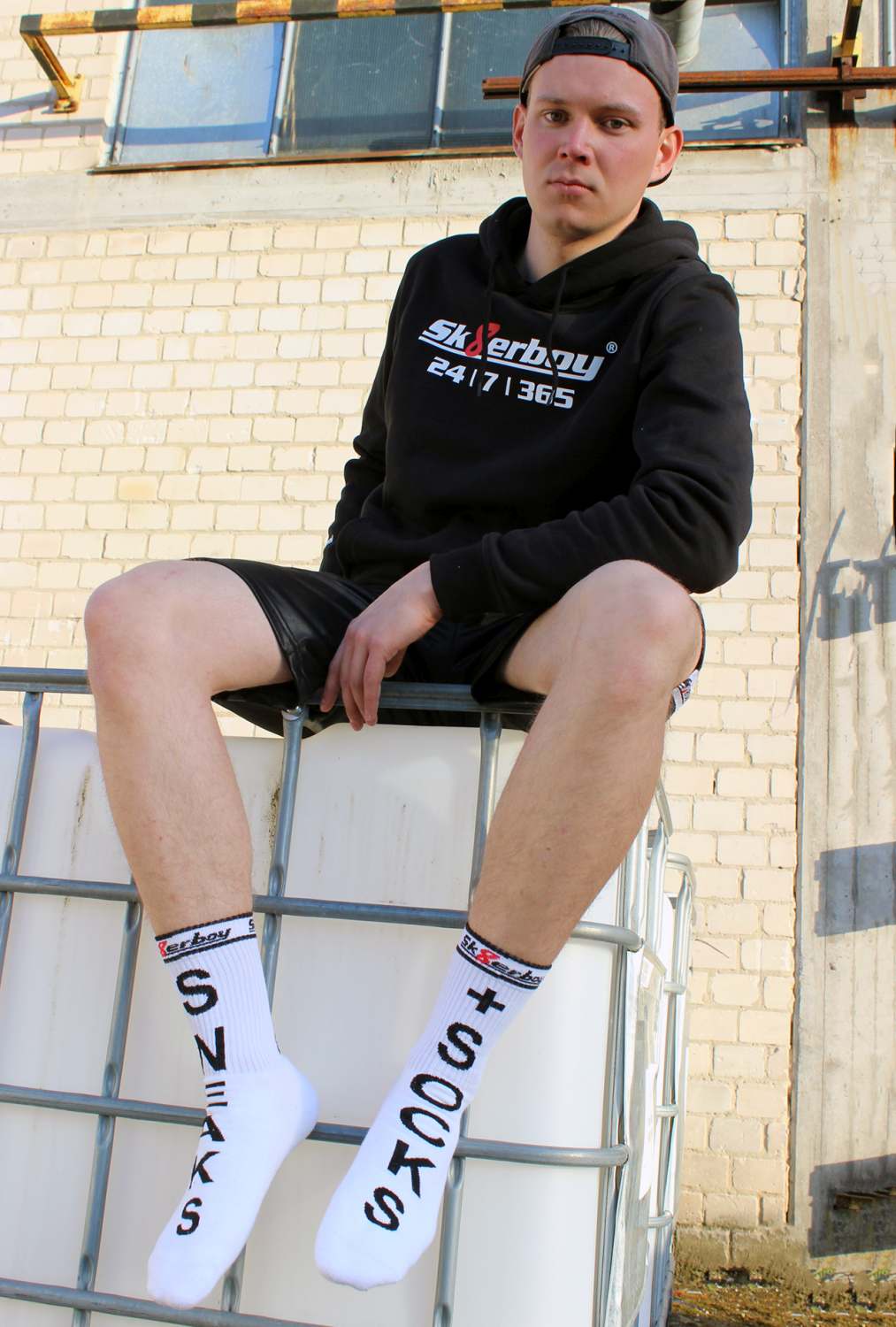 junger gay mit sneaks and socks socken von sk8erboy in weiß traegt einen schwarzen kaputzenpullover mi 365 7 24 aufdruck auf einem kontainer sitzend von vorne