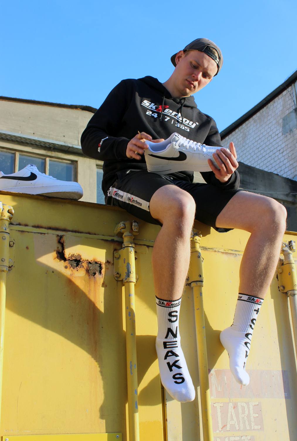 junger gay mit sneaks and socks socken von sk8erboy in weiß traegt einen schwarzen kaputzenpullover mi 365 7 24 aufdruck auf einem kontainer sitzend mit nike sneakern und shiny short unter blauem himmel