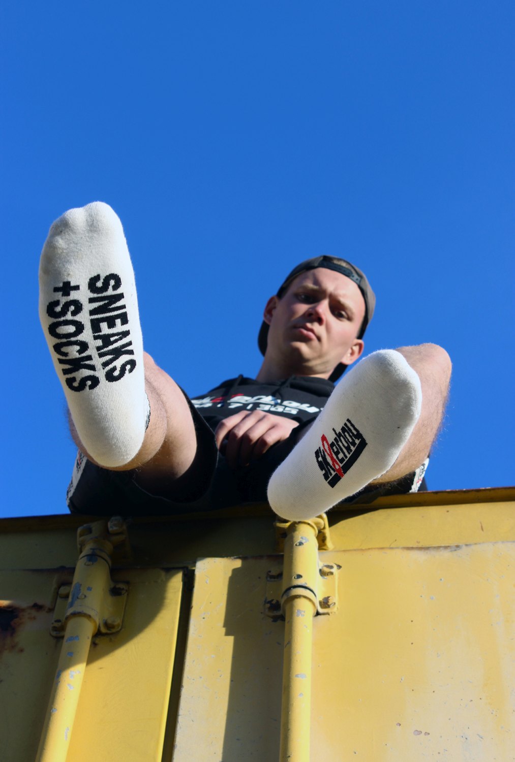 junger gay mit sneaks and socks socken von sk8erboy in weiß traegt einen schwarzen kaputzenpullover mi 365 7 24 aufdruck auf einem kontainer sitzend von unten mit blauem himmel