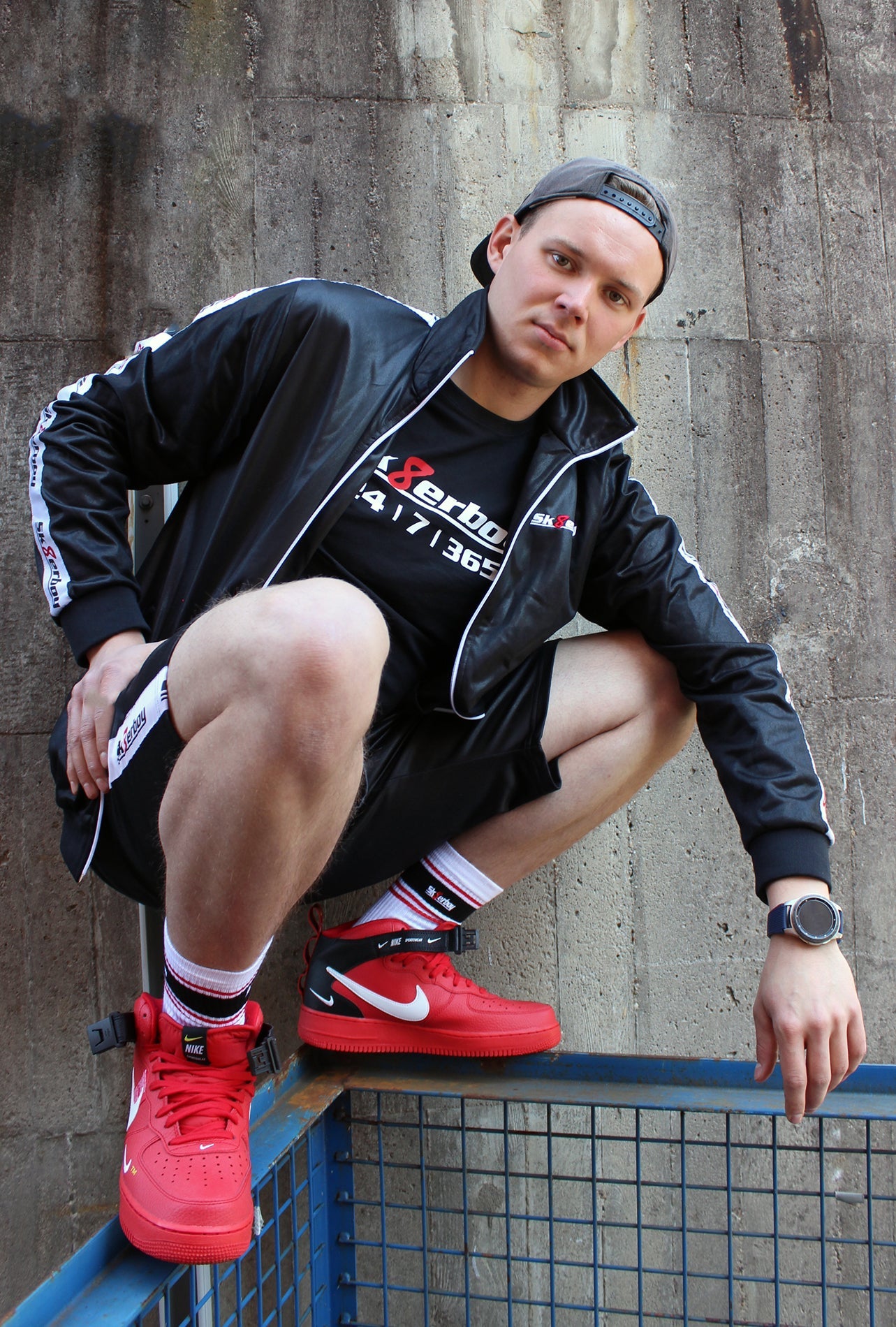 ein boy kerl bengel traegt ein sk8erboy t-shirt shirt in schwarz baumwolle cotton mit einem sk8erboy aufdruck mit weisser schrift und 8 acht in rot mit 24 7 365 fetter aufdruck weiter traegt er eine mund nasen bedenkung maske sk8erboy mit aufdruck dann hat er auf dem kopf eine basecap baseball seine graue jogger spant in der hose von nike air max und im hintergrund ein lost place alte fabrik